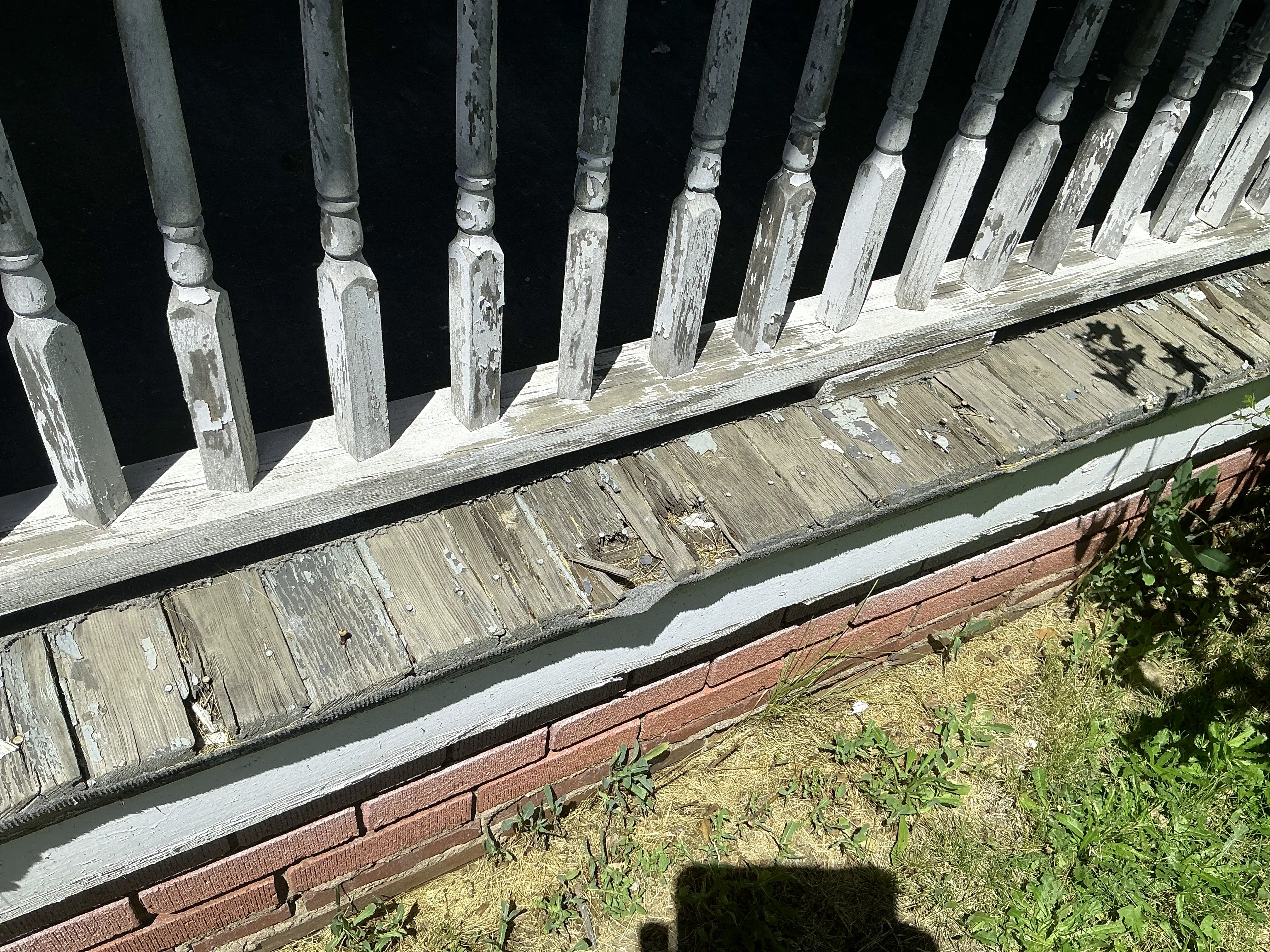 Weathered wood rot and peeling paint on porch floorboards below railing balusters 2026-08-10