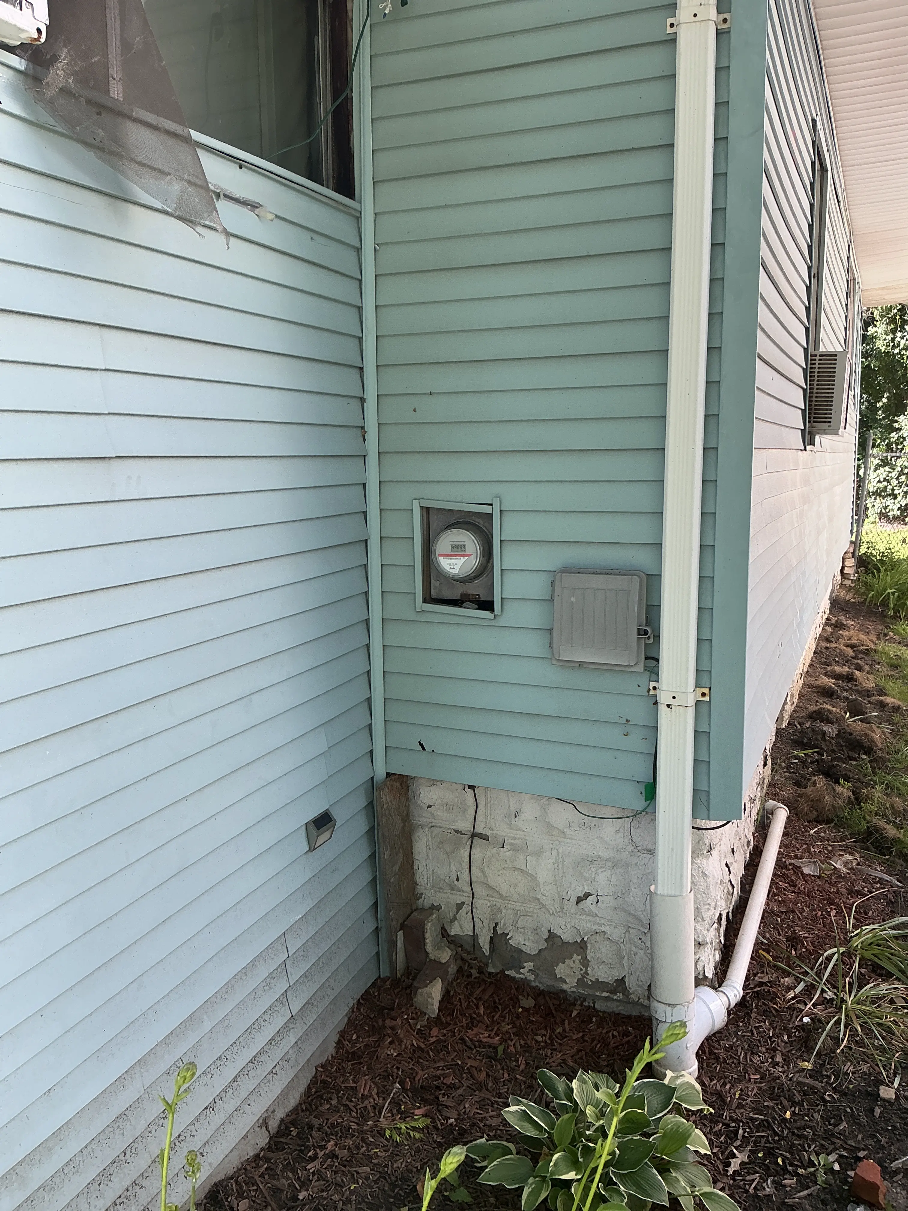 Deteriorated foundation parging coat along exterior home wall and garden bed. 2026-07-08