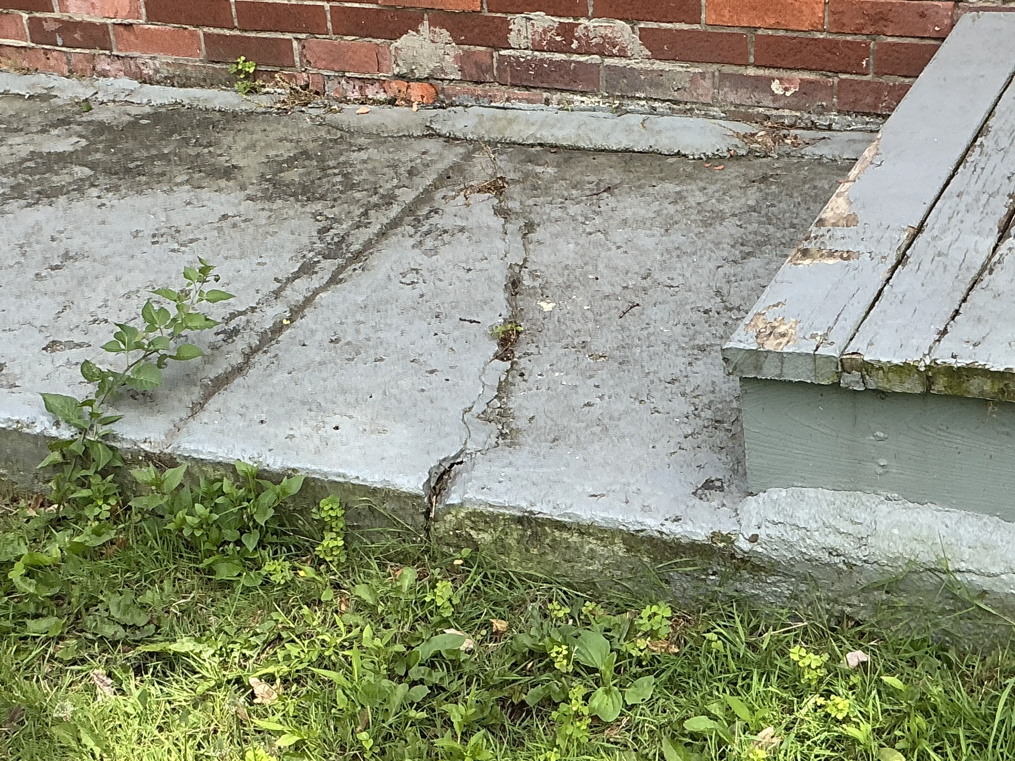 Cracked concrete walkway next to exterior wooden steps and brick wall 2026-07-18
