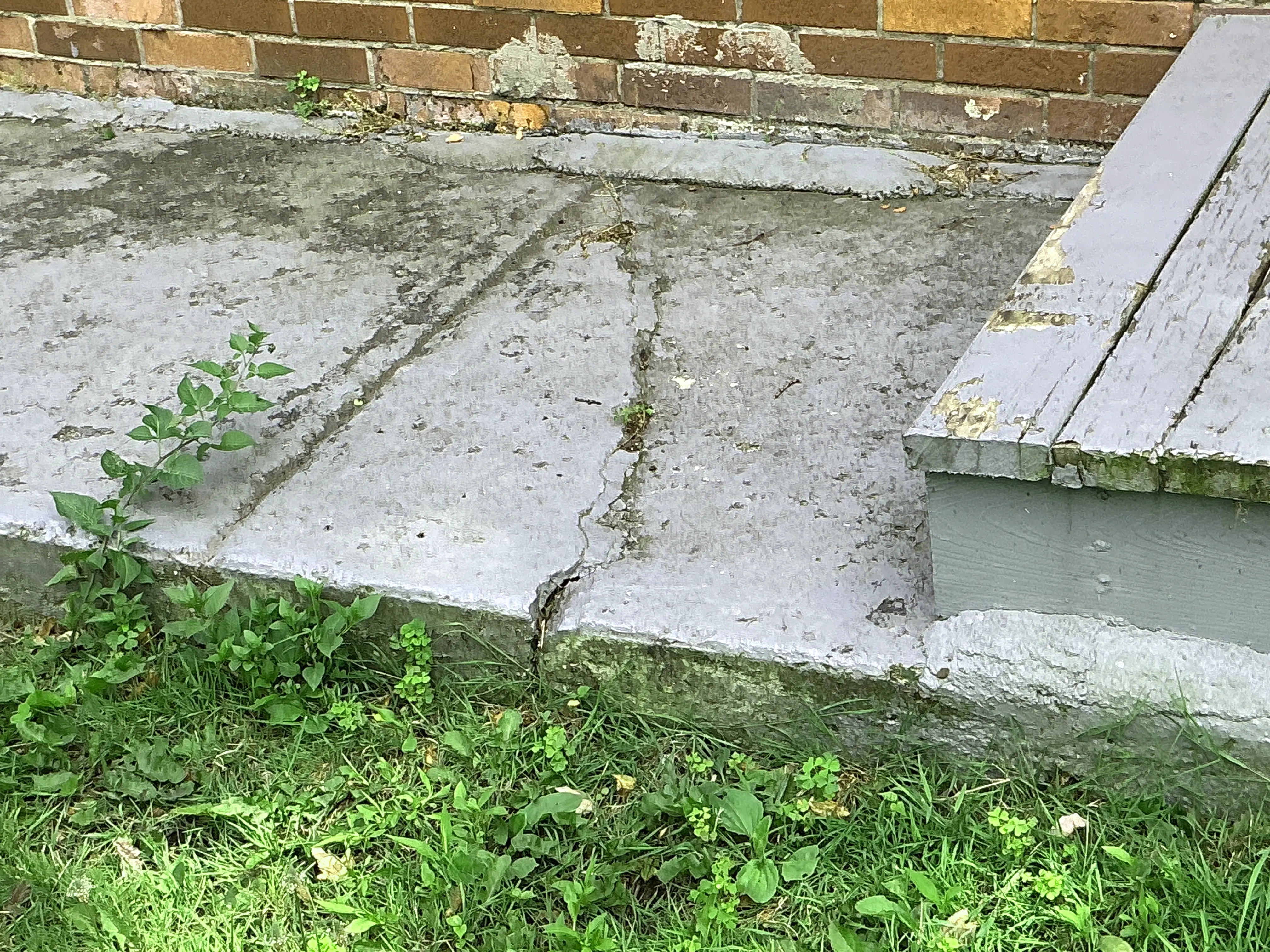 Cracked concrete walkway next to exterior wooden steps and brick wall 2026-07-05