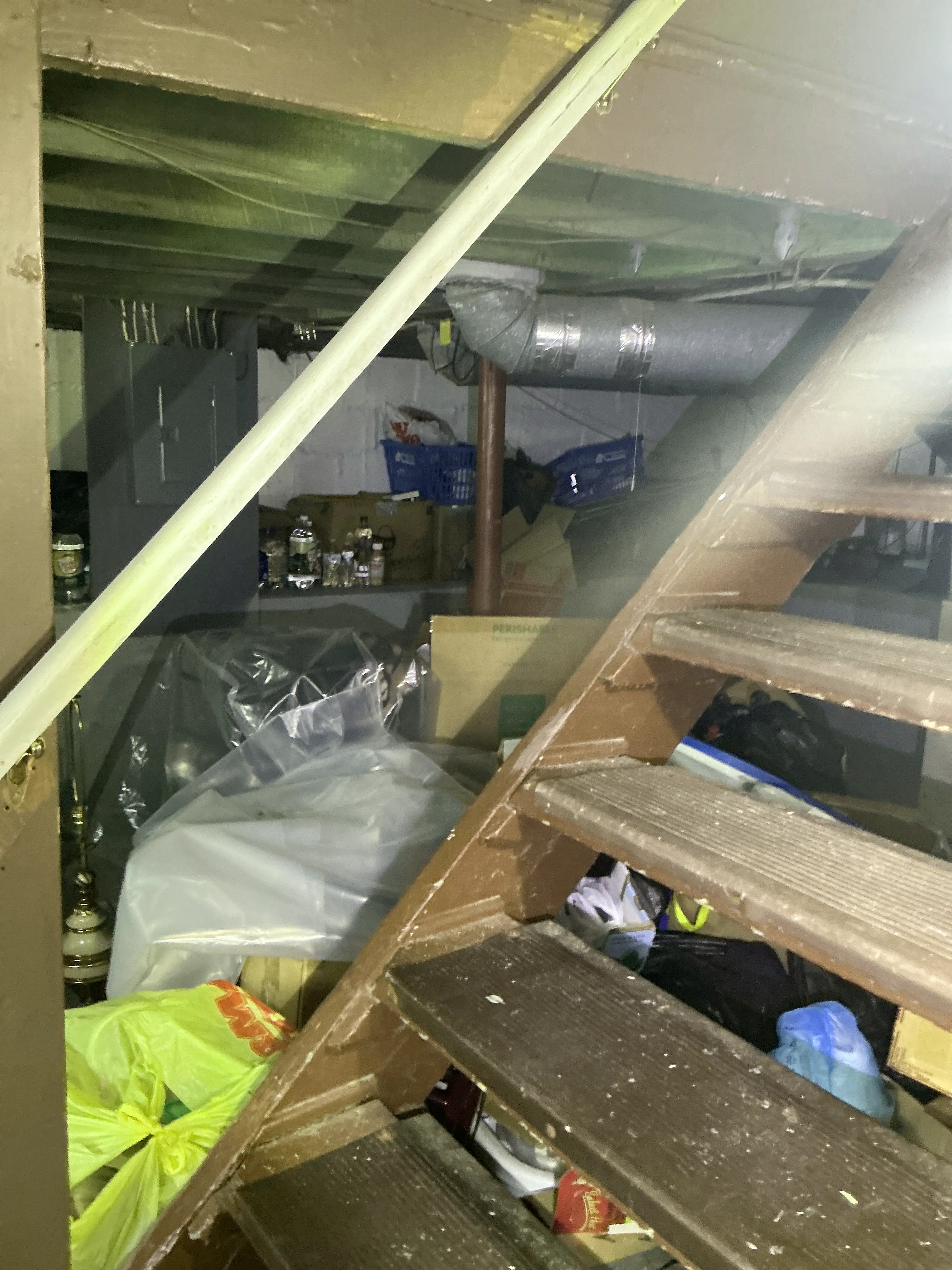 Cluttered storage area under wooden basement stairs filled with boxes, plastic bags, and visible ductwork 2026-07-20
