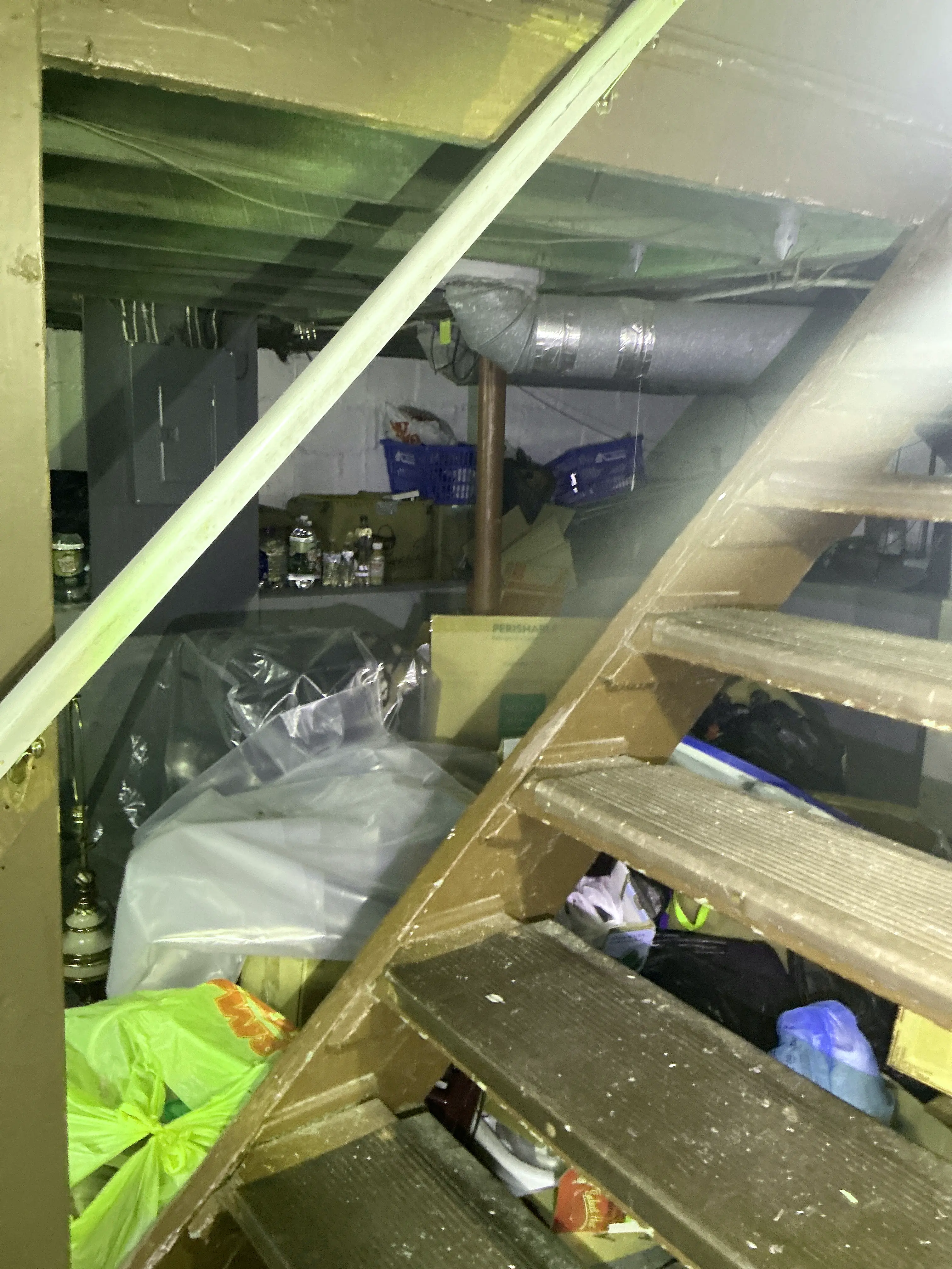 Cluttered storage area under wooden basement stairs filled with boxes, plastic bags, and visible ductwork 2026-07-24