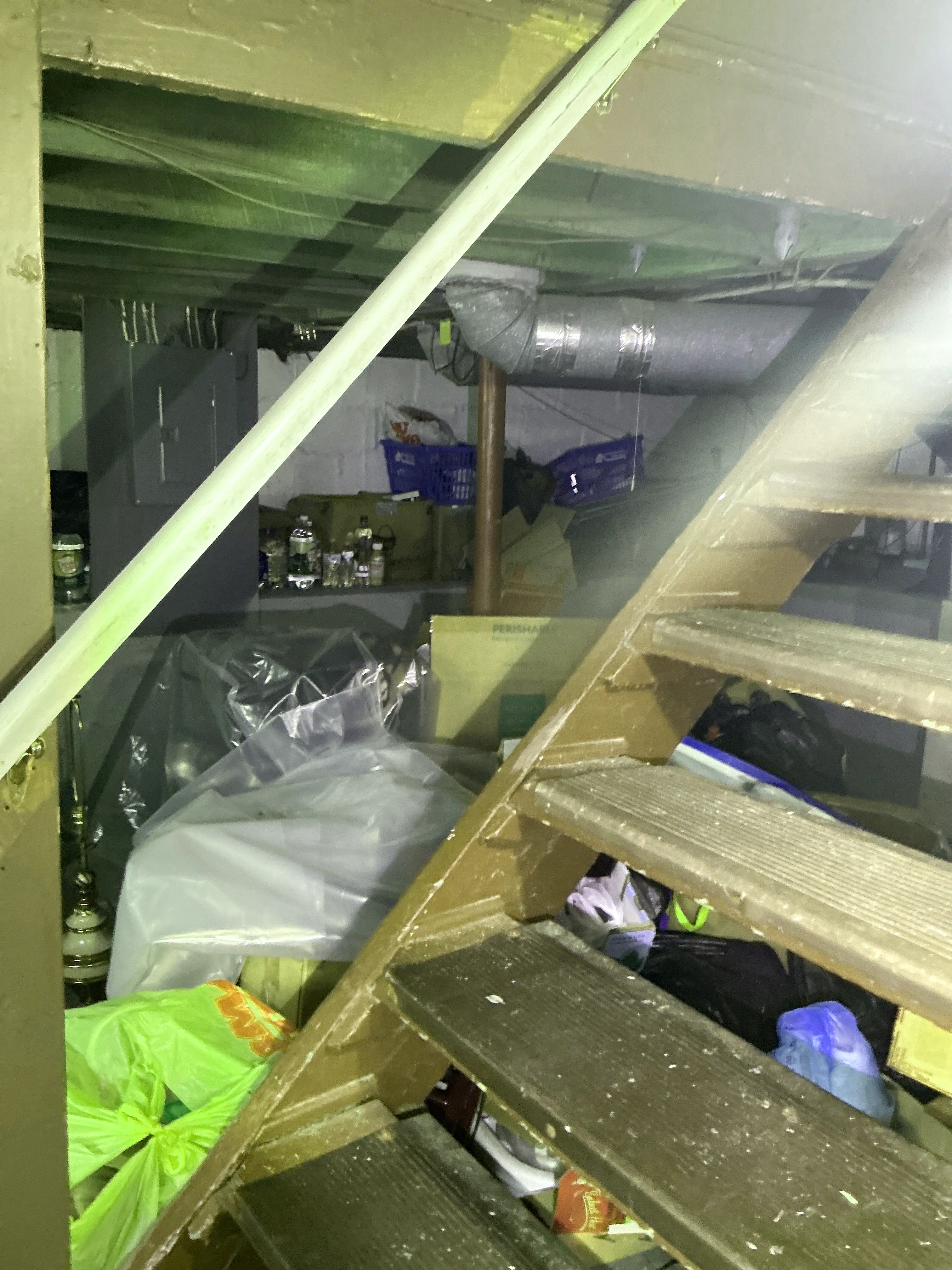 Cluttered storage area under wooden basement stairs filled with boxes, plastic bags, and visible ductwork 2026-08-02