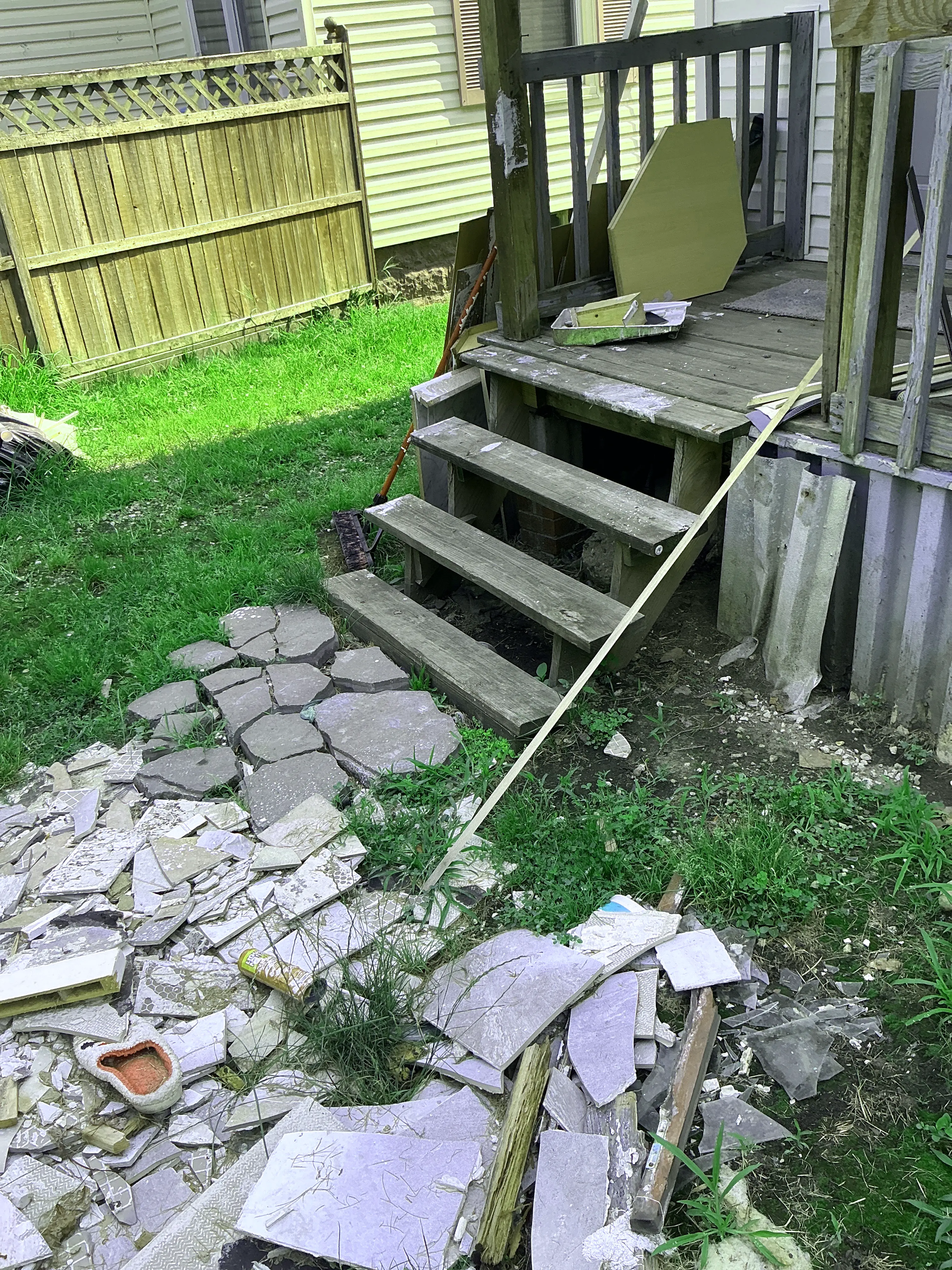 Cluttered yard with broken tiles and unsafe weathered wooden porch stairs. 2026-07-06