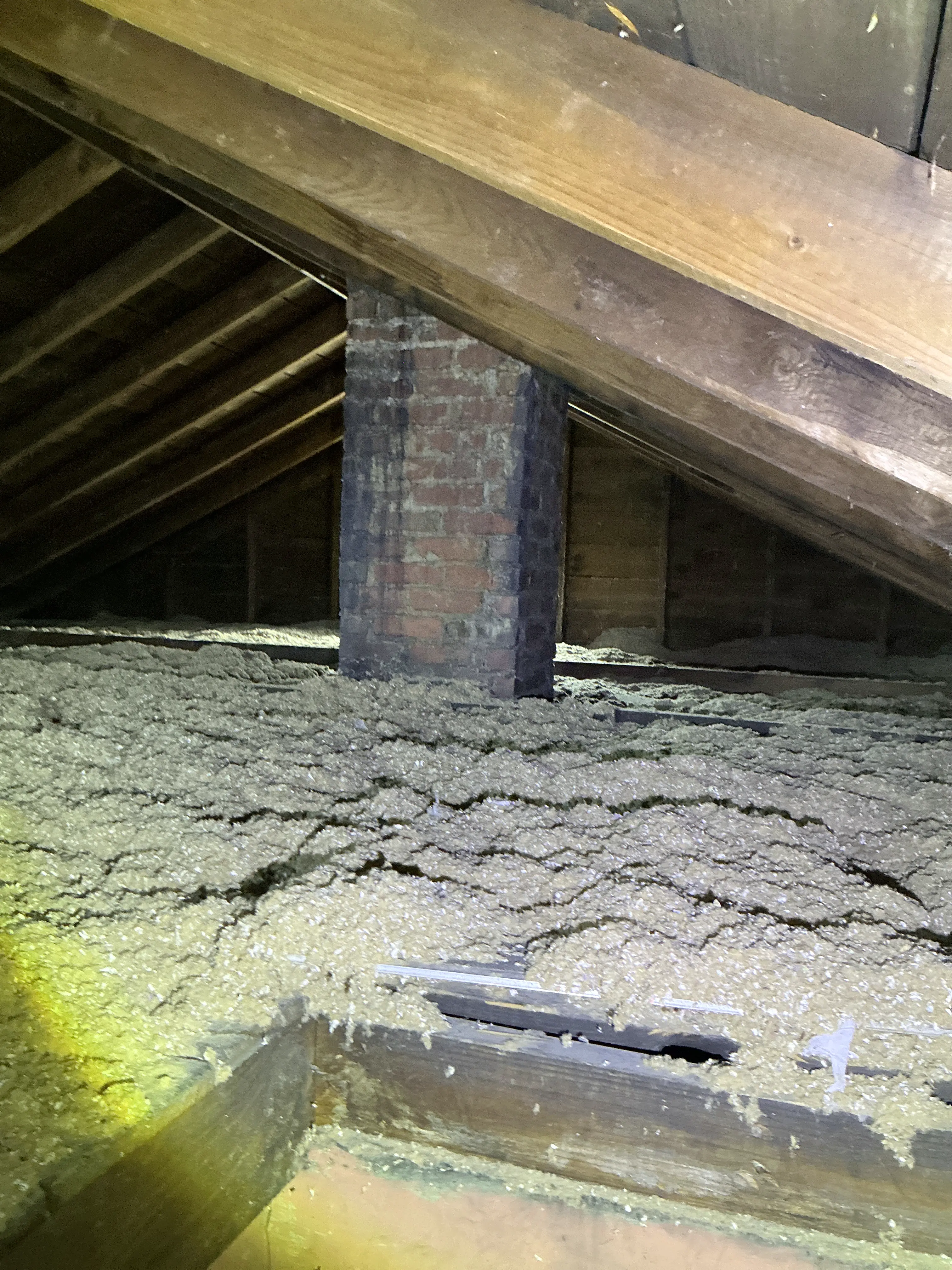 Brick chimney stack running through an attic space with blown-in insulation and exposed wood roof rafters. 2026-08-15