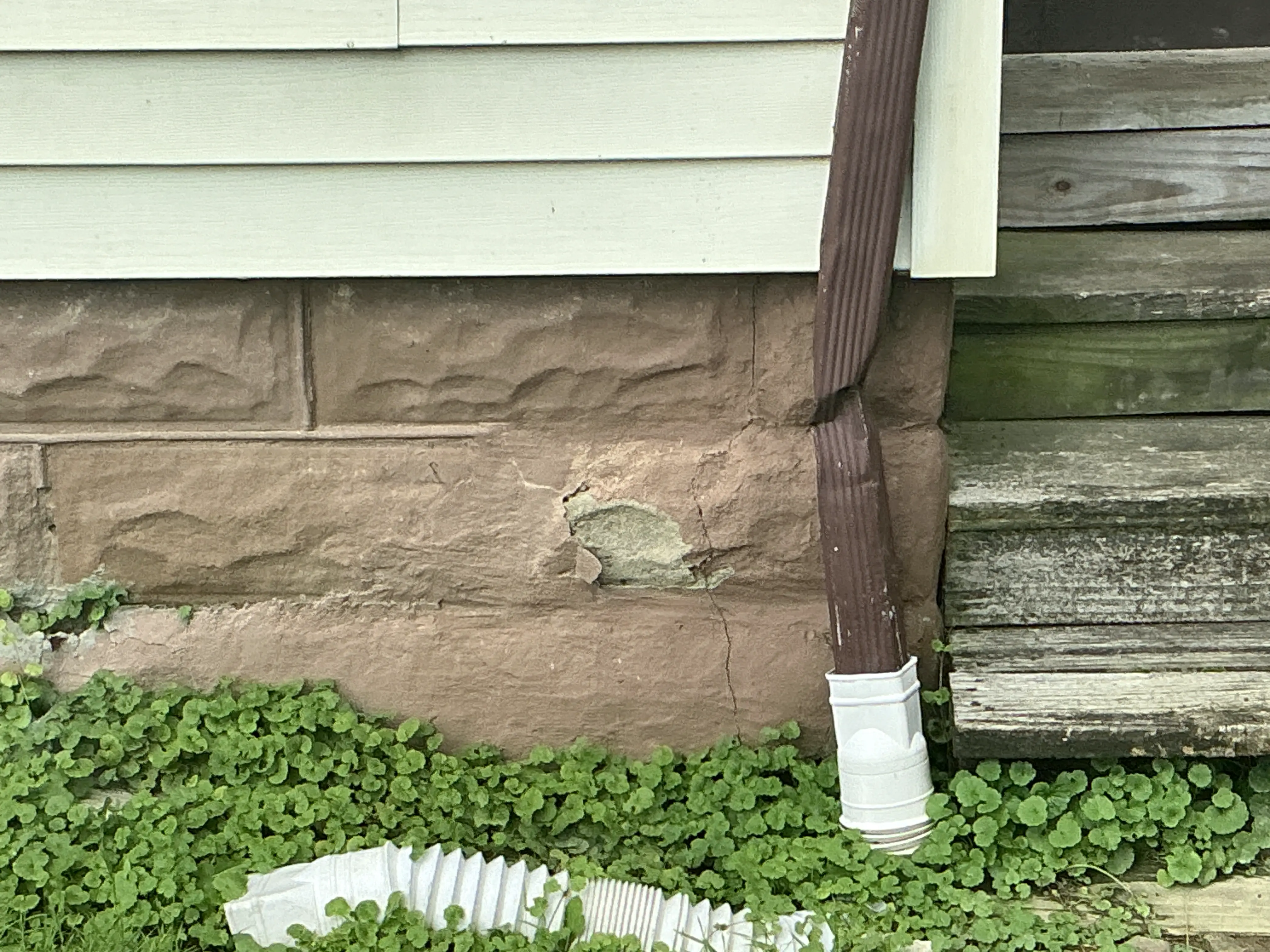 Cracked and spalling masonry foundation wall next to a crushed brown downspout. 2026-07-31