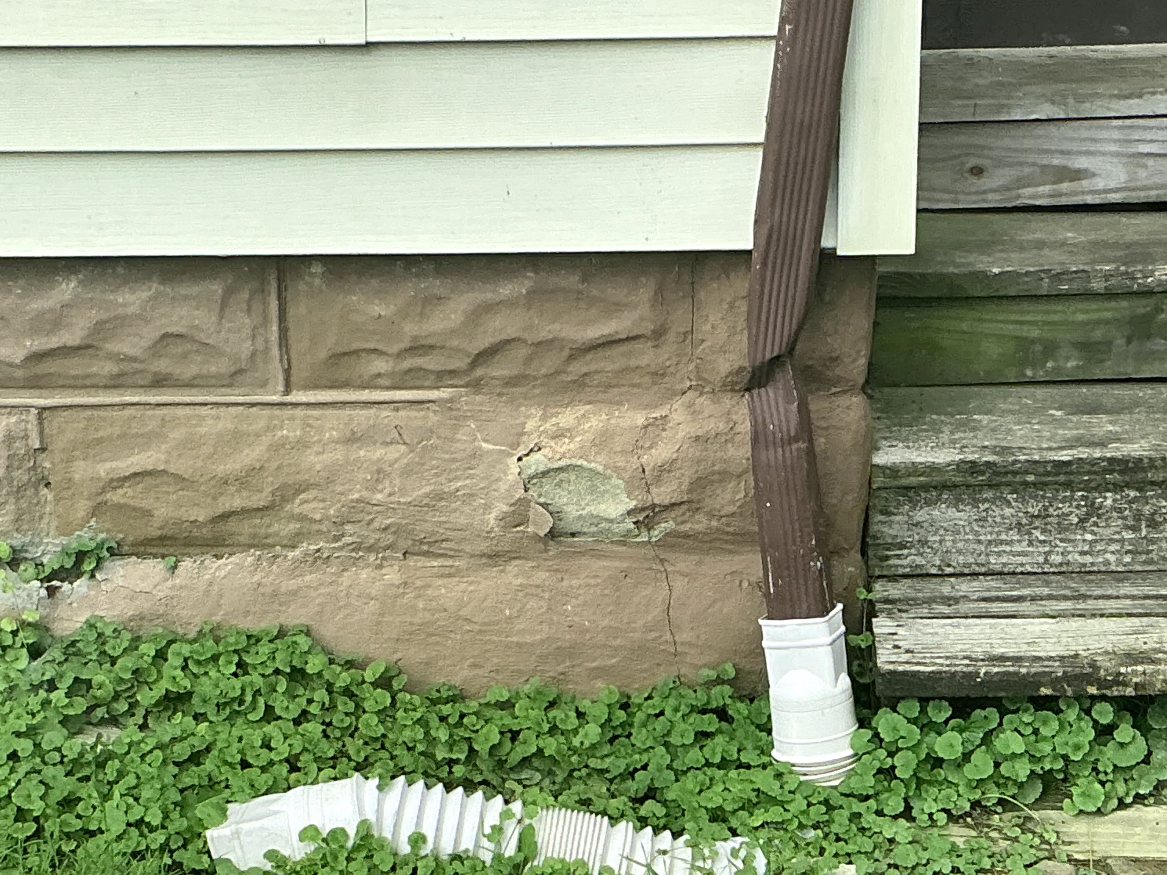 Cracked and spalling masonry foundation wall next to a crushed brown downspout. 2026-07-27