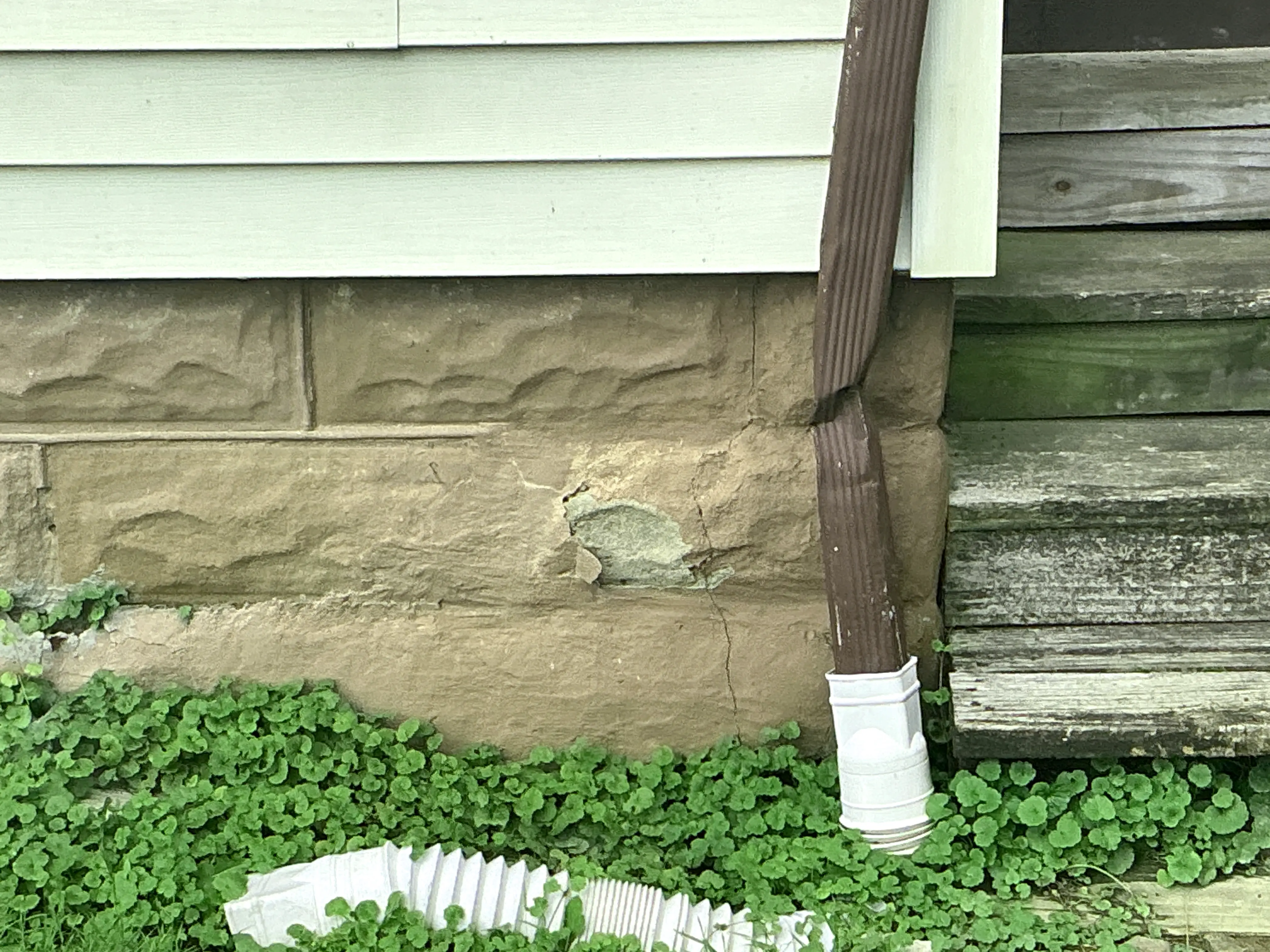 Cracked and spalling masonry foundation wall next to a crushed brown downspout. 2026-08-26
