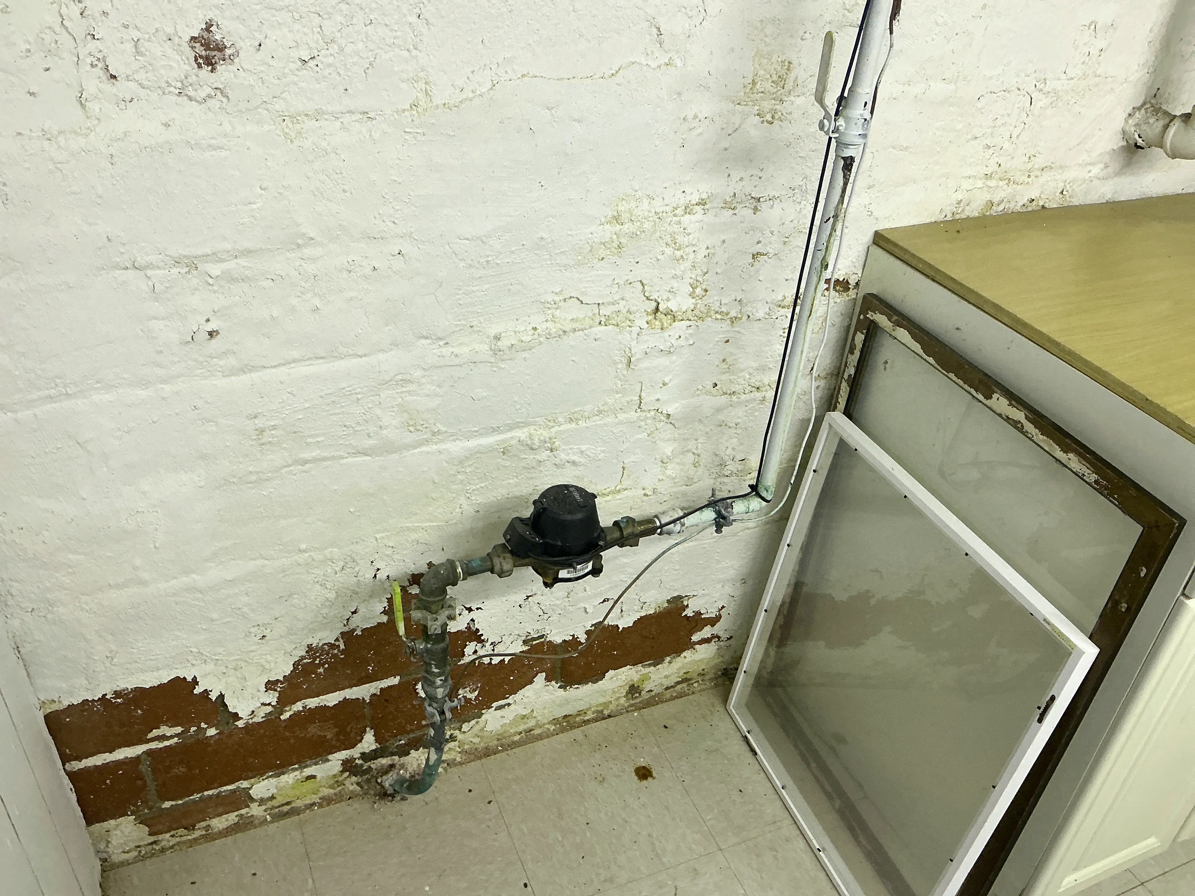 Water meter and shutoff valve against a painted brick basement wall with peeling paint 2026-08-18